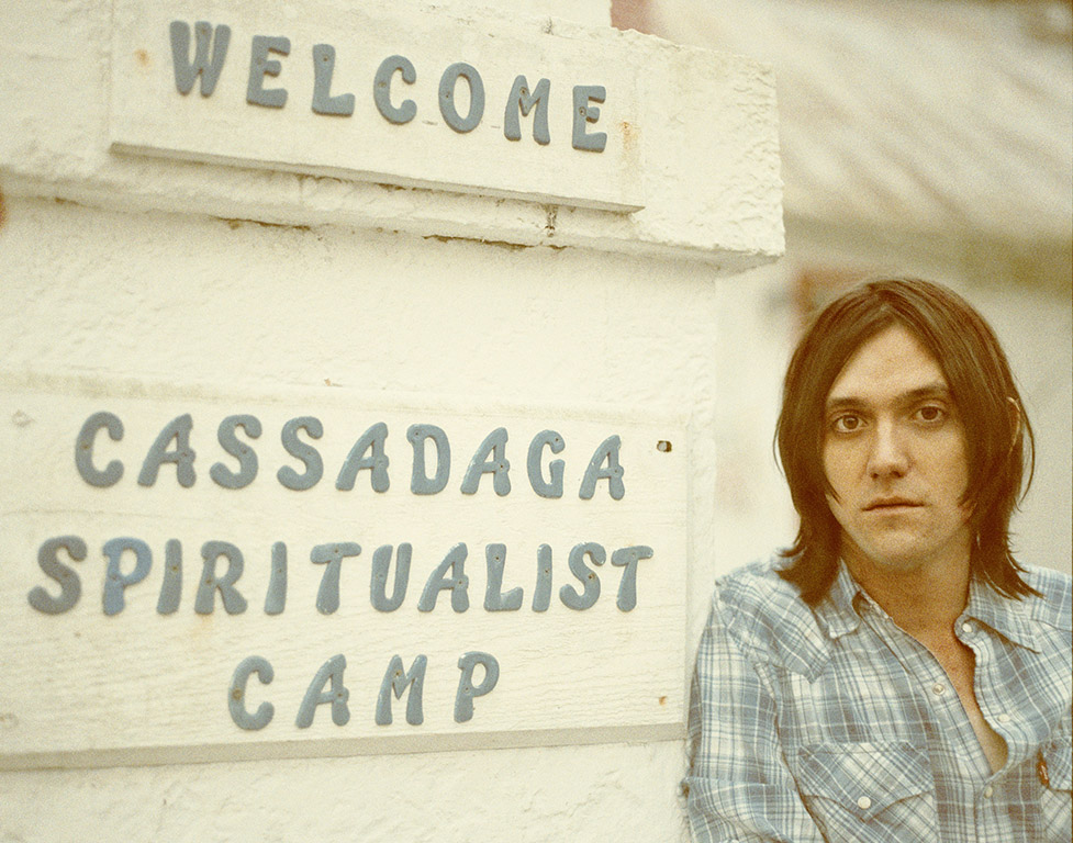 Connor Oberst leaning against a white building with a sign in blue writing that says in Welcome, Cassadaga Spiritualist Camp
